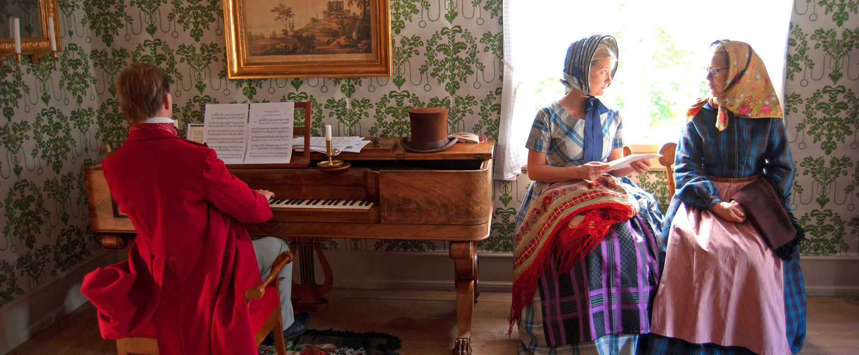 Kläder i hus & gårdar En man spelar klaviatur, två kvinnor sitter bredvid och samtalar.