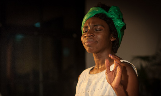 A close up of Apphia Campbell who stands with her eyes closed and palm raised. She is wearing a green headscarf.