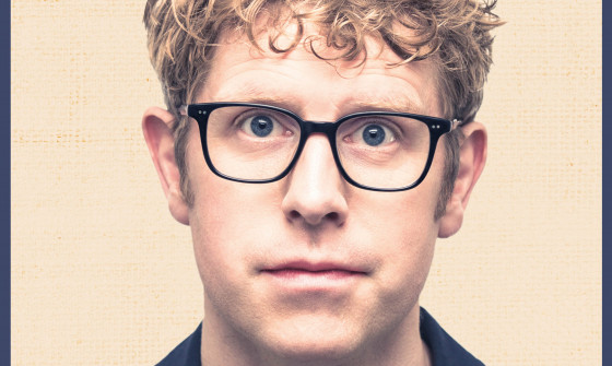 A close up of Josh Widdicombe with curly blonde hair, black glasses and a stripey red t-shirt.