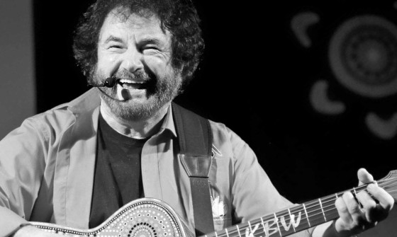 Kevin Bloody Wilson laughs on stage holding his guitar in its aboriginal design. The photo is black and white and at an angle.