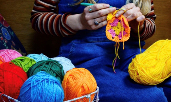 Colourful balls of yarn take the forground of the photo, with a woman crocheting a circular pattern in the background with yellow yarn.