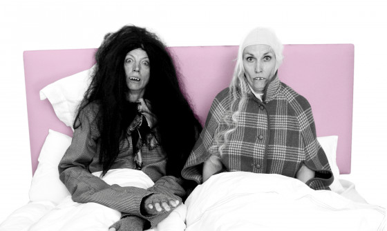 The two leads of Enter the Dragons sit in a bed with fake teeth and wigs. The photo is in black and white, with the headboard of the bed in a pastel pink.