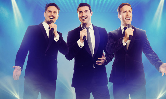 The three members of Blake stand on stage in suits with microphones, looking beyond the camera on a blue background with spotlights.