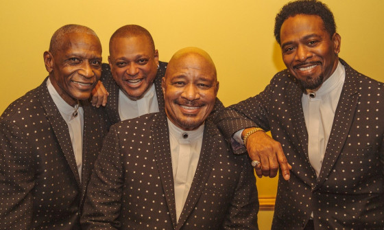 The four members of The Stylistics stand in front of a yellow background in matching brown suits with white embellishment.