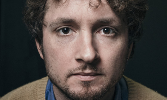 A close up of Sam Amidon who looks directly into the camera. He has dark brown curly hair, and is wearing a blue shirt with a brown jumper on top. The background is black.