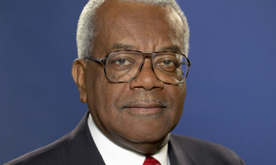 Sir Trevor Mc Donald is facing the camera smiling. He is wearing a black suit with a red tie. There is a dark blue wall behind him