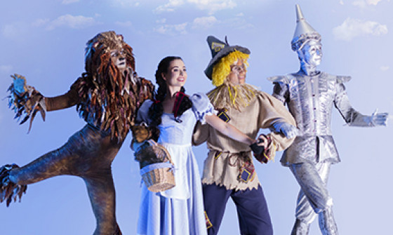 The Lion, Dorothy, Scarecrow, and Tinman are all linking arms as they skip across a green hill