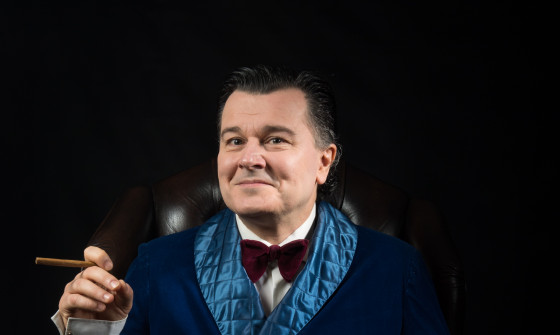 A man is sat in a Professor armchair. He is wearing a navy smoking jacket which has a light blue quilted trim. His right elbow rests on the chair arm, in his hand he has a cigar. His left hand is holding a glass of red wine. The background is black.