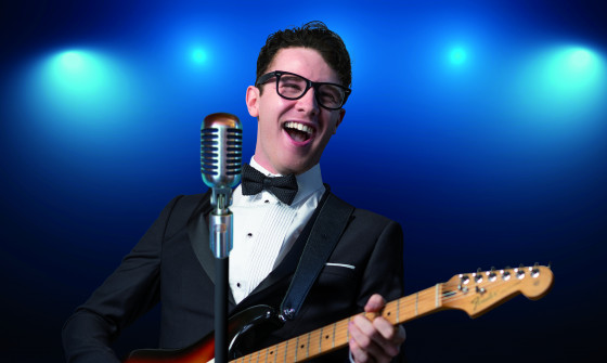 A mid-shot of Buddy Holly playing his electric guitar. He is wearing a black tuxedo. Blue stage lights shine behind him