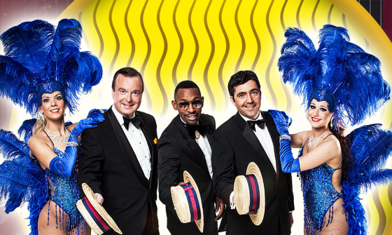 A full shot of three male members of the cast. They are all wearing a black tuxedo and are holding out boater hats in front of them. At either side of the men on the end, stands a lady dressed in a blue burlesque feather costumes.