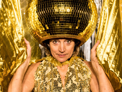 A woman with a golden disco ball on her head stands in front of a gold curtain