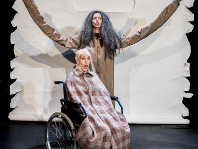 A woman with very long arms stands behind a character in a wheelchair who sits beneath a blanket
