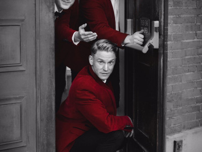 The members of Bye Bye Baby poke their heads out of a door. The image is in black and white with just suits in red.