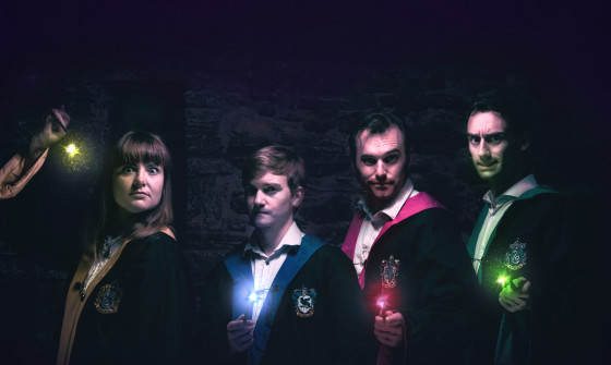 Four cloaked Hogwarts students stand in front of a Hogwarts shadow with wands