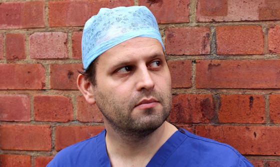 Adam Kay is dressed in blue hospital scrubs and is standing against a brick wall