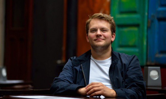 Mid shot of Valeriy Sokolov sat down. He is wearing a navy jacket with a white t shirt