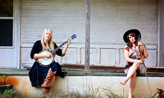 The two members of Local Honeys sit on a rustic porch. One holds a banjo, and the other holds a violin.