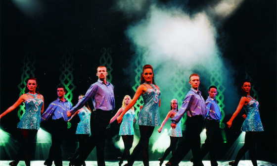 A dance troupe perform on stage in traditional Irish dance attire. They all stand with one leg behind them with a pointed toe, and their arms behind them also.