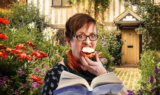 Helen Wood stands in front of a quaint cottage surrounded by flowers. She is eating a scone and reading a National Trust guide. She has cream on her nose.