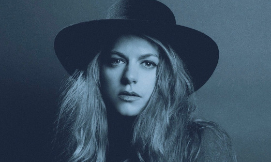 A close up of Jo Harman who looks at the camera, wearing a gambler-style hat. She has long wavy hair that frames her face.