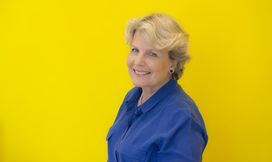 Sandi Toksvig smiles at the camera. She is wearing a royal blue shirt which contrasts the striking yellow backdrop.