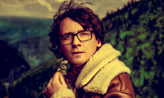 Ed Byrne stands in a highland setting surrounded by trees, in an brown aviator jacket. He holds a shrub to his face.