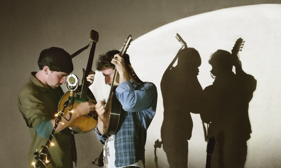 Ye Vagabonds stand together with their banjos in the air facing eachother. They shadow is cast onto a wall behind them,
