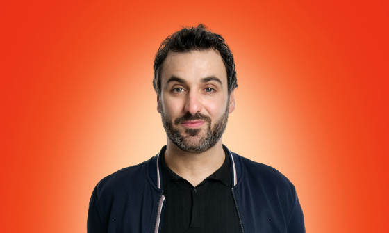 Patrick Monahan stands casually in front of an orange background. He smiles at the camera.