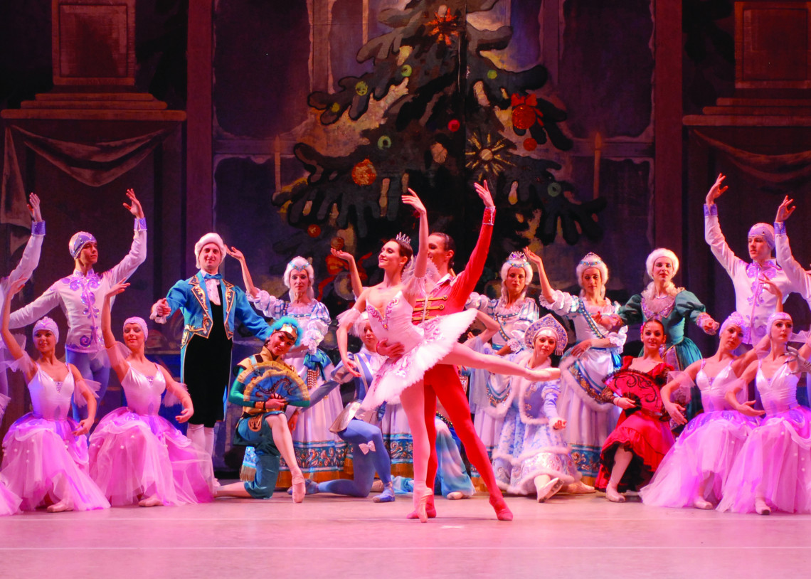 The Nutcracker Prince dances with the Sugar Plum Fairy. Other characters fill the background in front of a large Christmas tree.