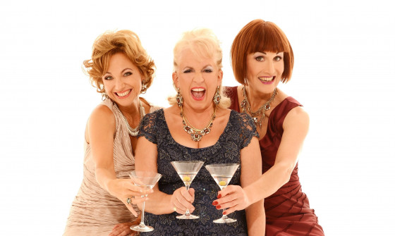 Three women stand in front of a white background cheersing martini glasses.