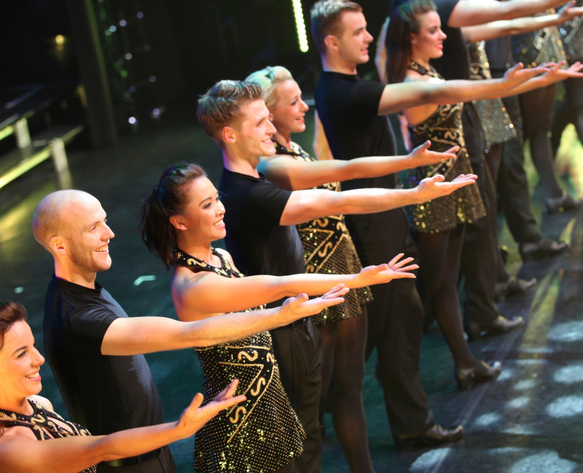 A line of dancers extend their arms in unison, smiling