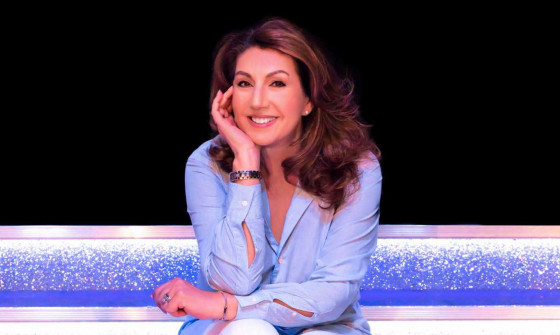 Jane Mc Donald sit on silver steps resting her head in her hand. She is smiling towards the camera