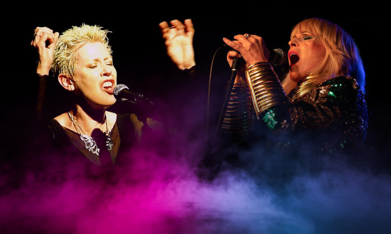 Toyah and Hazel O'Connor stand behind coloured smoke on screen both singing into the microphone.