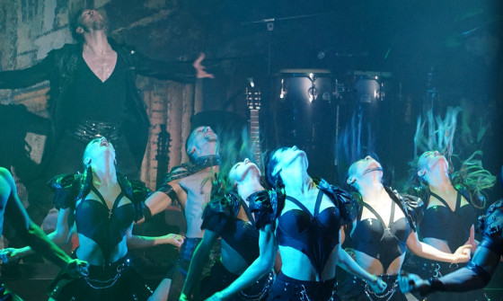 A close up of dancers in black leather costumers dance under a green light.