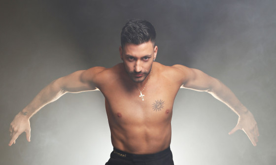 A shirtless Giovanni stands backlit pulling a dancing pose.