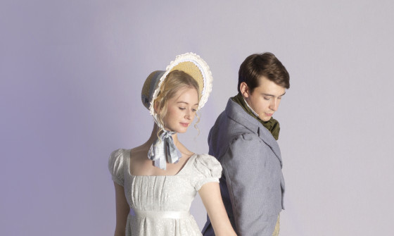 A costumed male and female stand next to eachother with their hands touching in ballet poise.