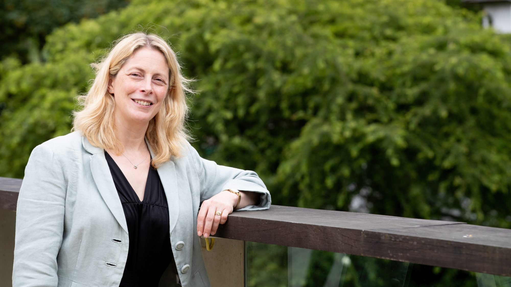 A woman with blonde hair is standing on a bridge, with blurred trees in the background.