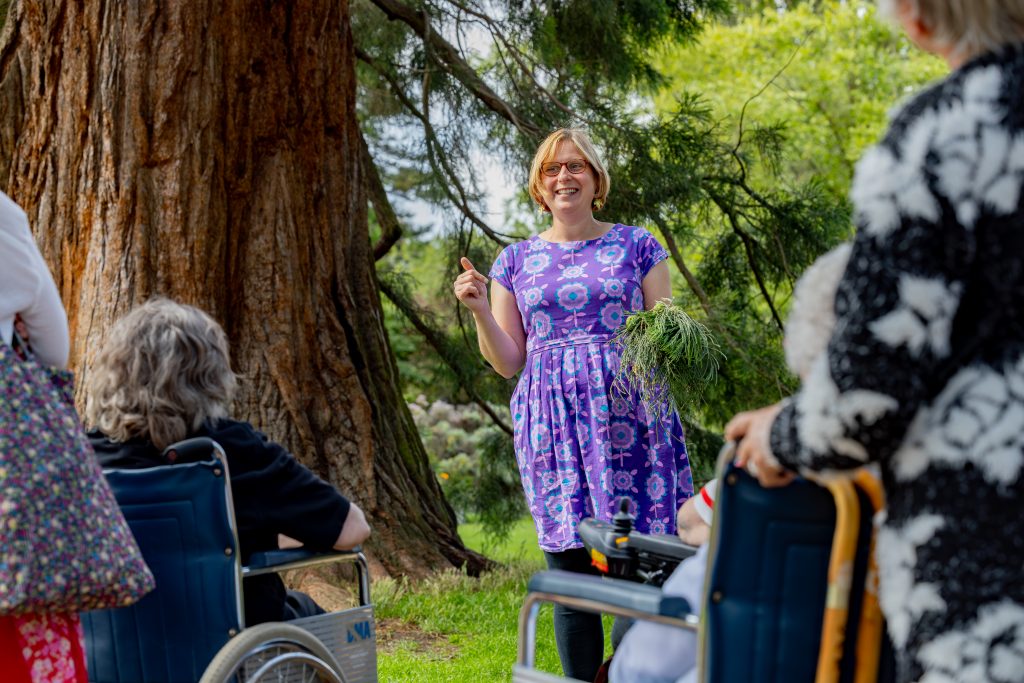 Woman in purple dress holding some foliage talking to a group of people in wheelchairs