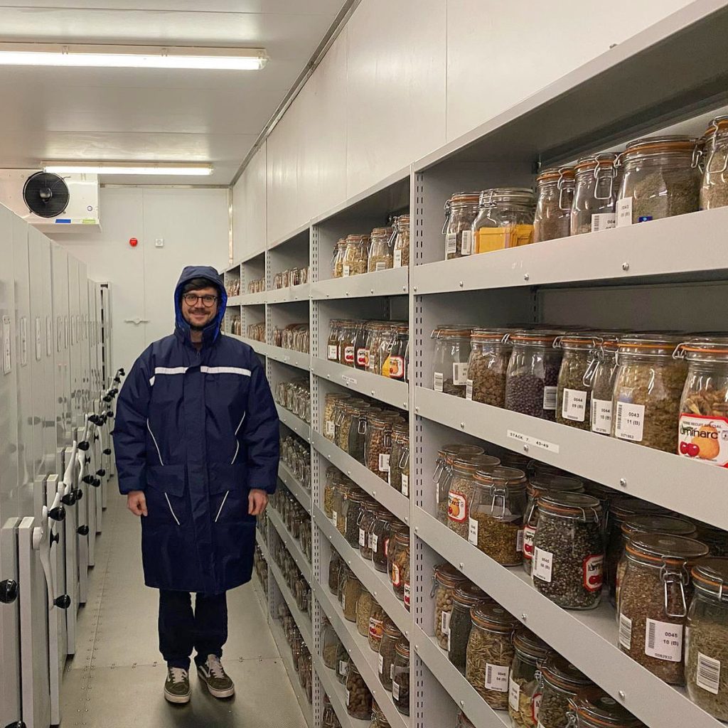 A man in a large blue coat smiles from within a seed bank, with rows of different seeds on shelves.