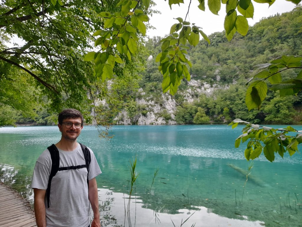 A man in a grey shirt smiles at the camera in front of a turquoise-watered lake.