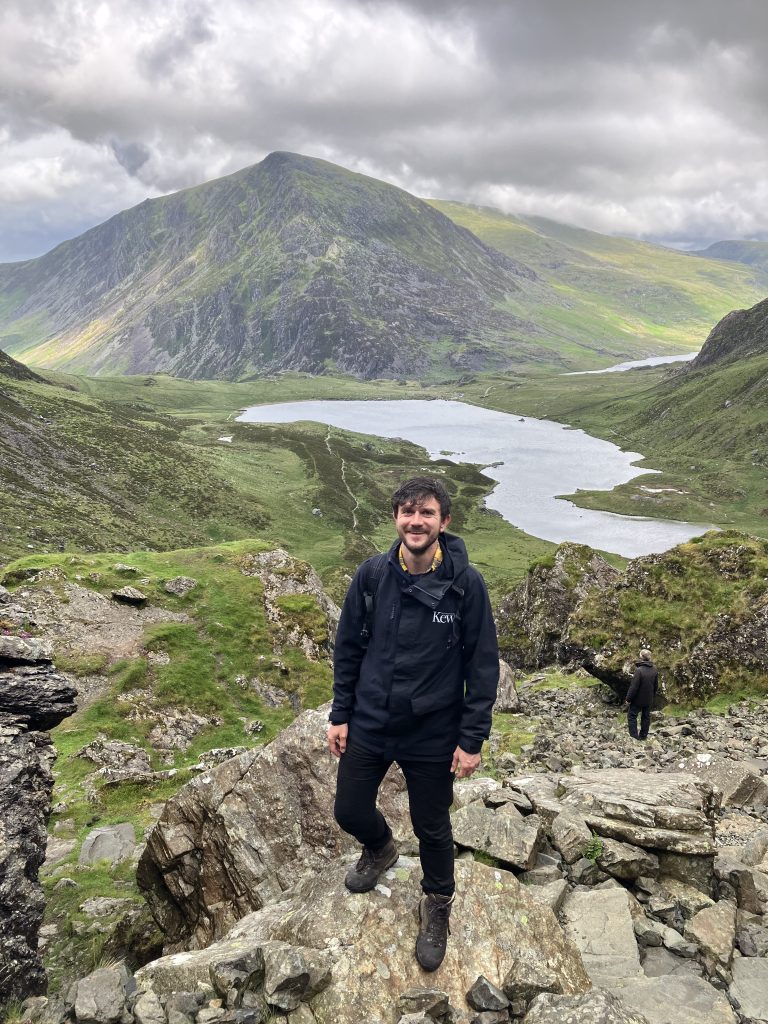 A man in black Kew-branded clothing stands in front of mountains and valleys.