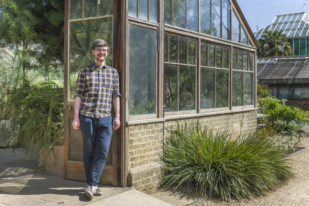 Matthew, in a blue yellow plaid shirt, smiles as he leans against the doors of the glasshouse.