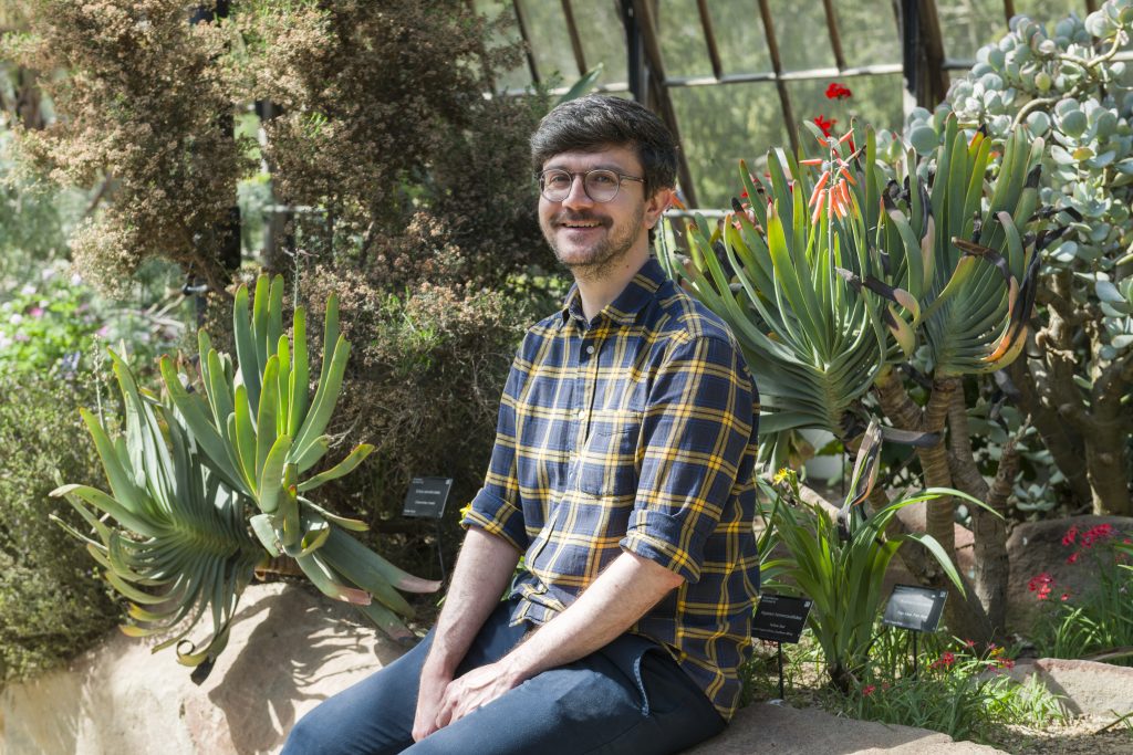 Matthew, in a blue yellow plaid shirt, smiles as he sits inside a glasshouse surrounded by plants.