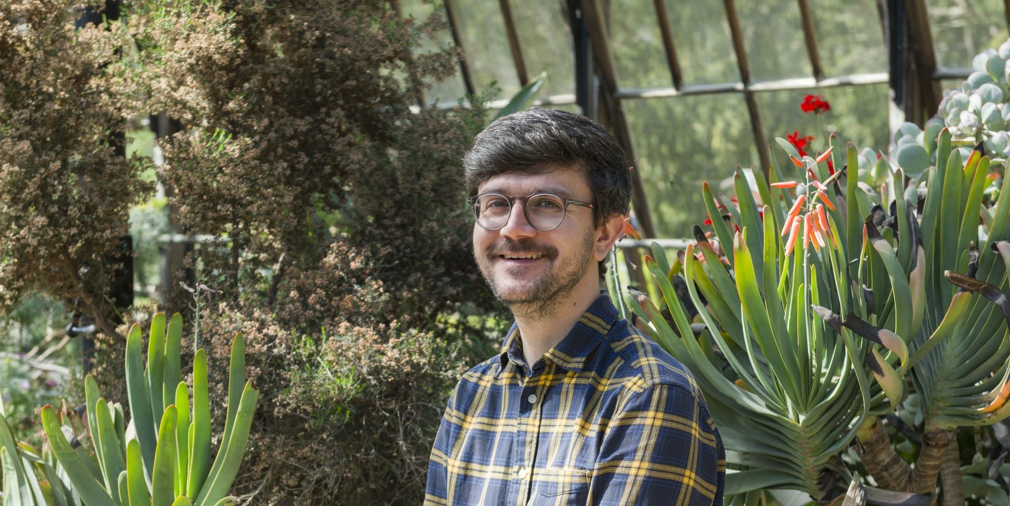 Matthew, in a blue yellow plaid shirt, smiles as he sits inside a glasshouse surrounded by plants.