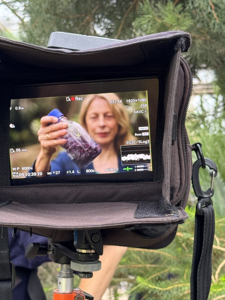 CUBG Director, Beverley Glover seen through a TV camera lens holding up a clear jar filled chopped with red cabbage
