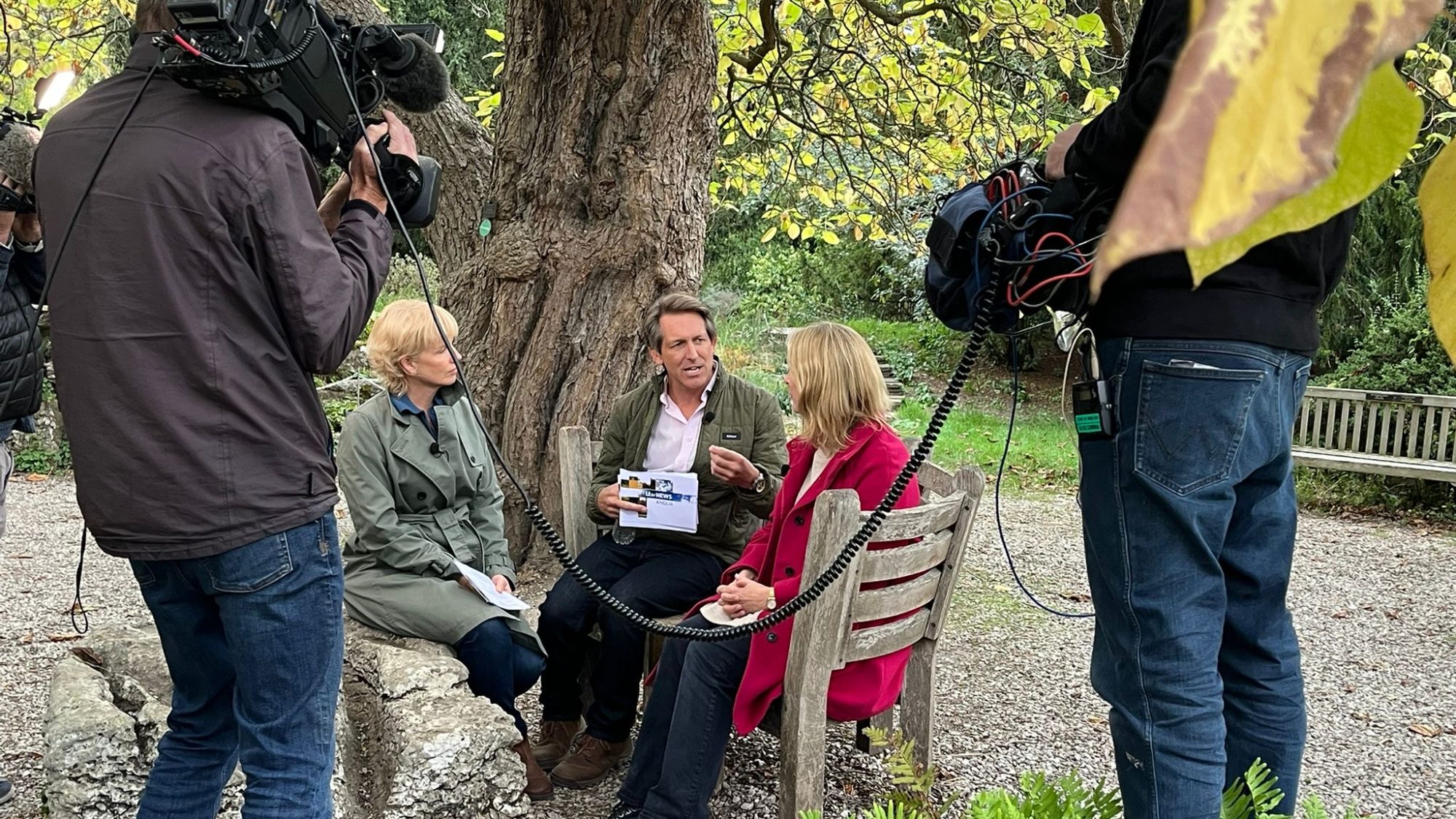 Three people sit talking on a bench under a tree while they are filmed by two other people.