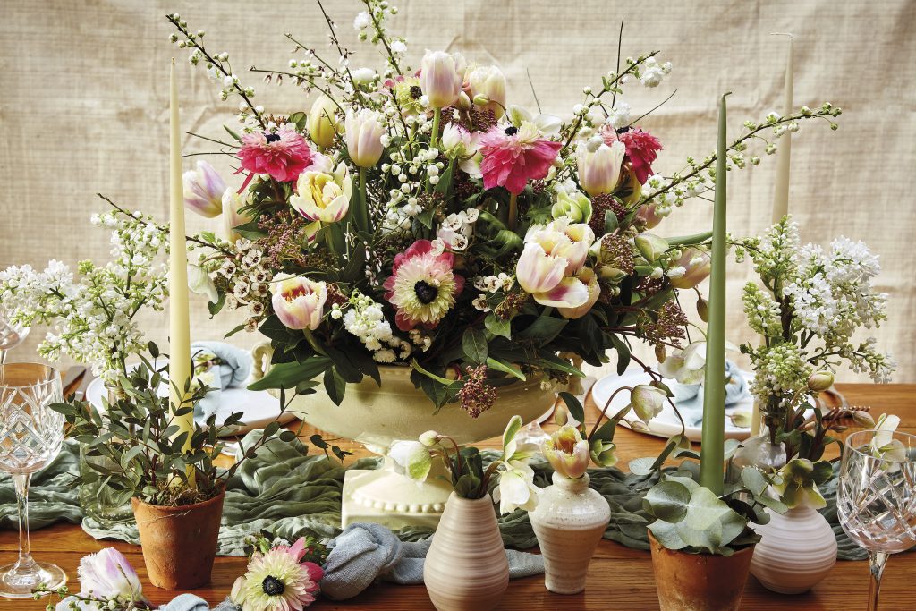 An centrepiece arrangement of pink and cream anemones on a table surrounded by candles and plates 