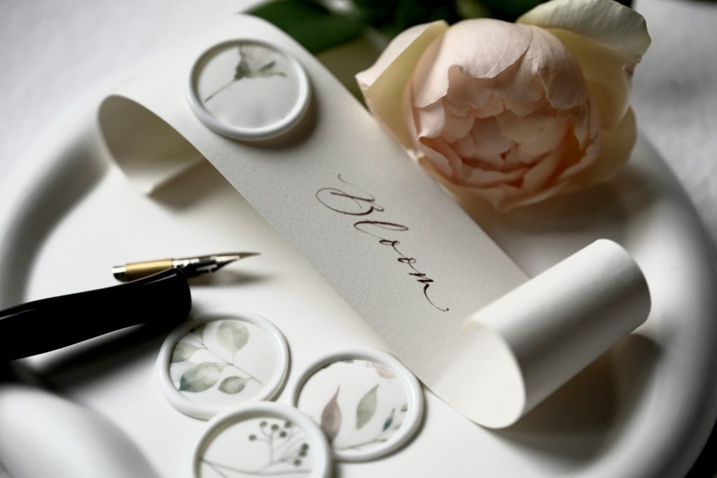 Small scroll of paper with the word 'Bloom' hand written on it, next to a rose and a pen