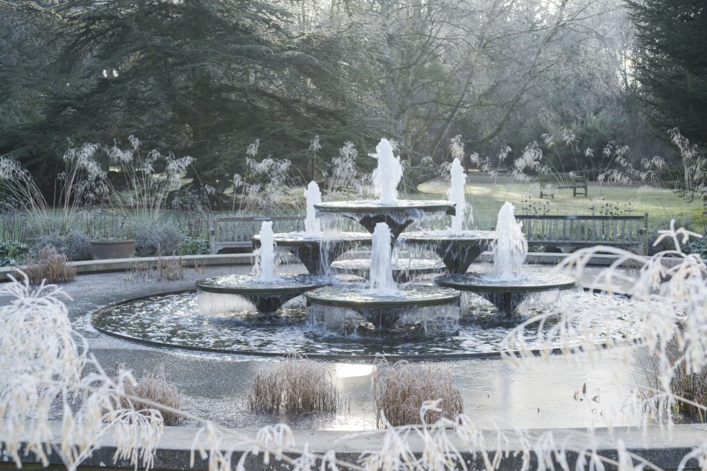 A fountain surrounded by frost covered trees, plants and shrubs.
