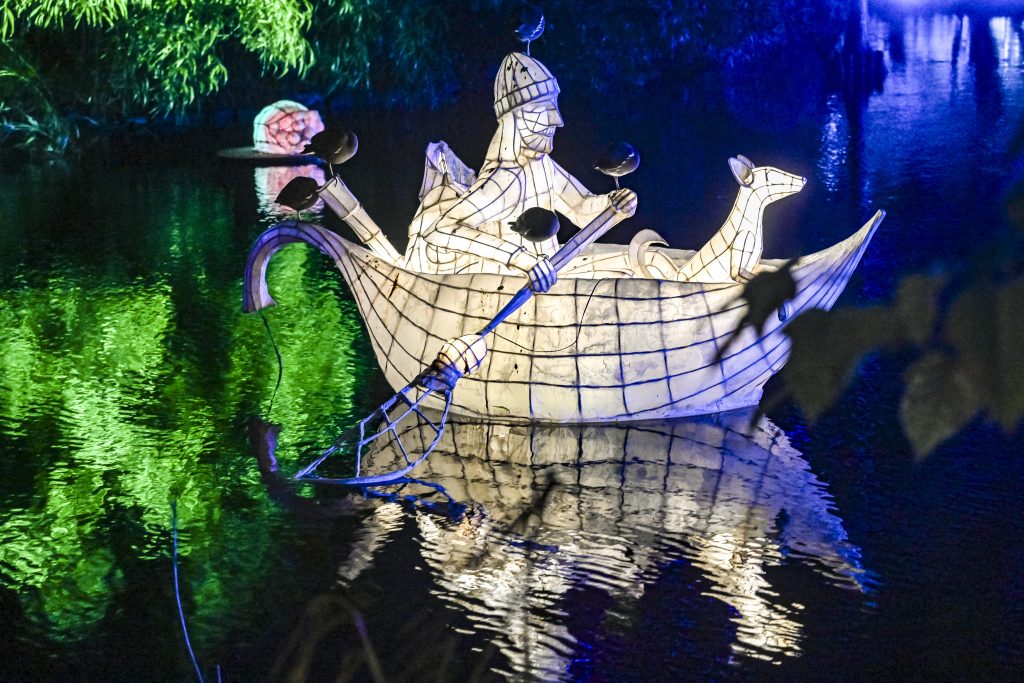 A three-dimensional sculpture of a person in a row boat is set on the lake. The sculpture is made of wire and a material that resembles papier-mâché, and is lit from within giving it a warm white glow. A sculpture of a dog made from the same material sits in the front of the boat, and the human figure holds an object shaped similarly to a net which is hooked towards the water, suggesting they may be scooping something from the lake. On the sculpture, five live moorhens are roosting - one on each of the person's hands, two towards the stern of the boat, and one perched on the figure's head. In the background is another sculpture of the same materials - this time a seed lit in a rainbow of colours, The lake reflects green light to the left of the image, and blue light to the right of the image. 